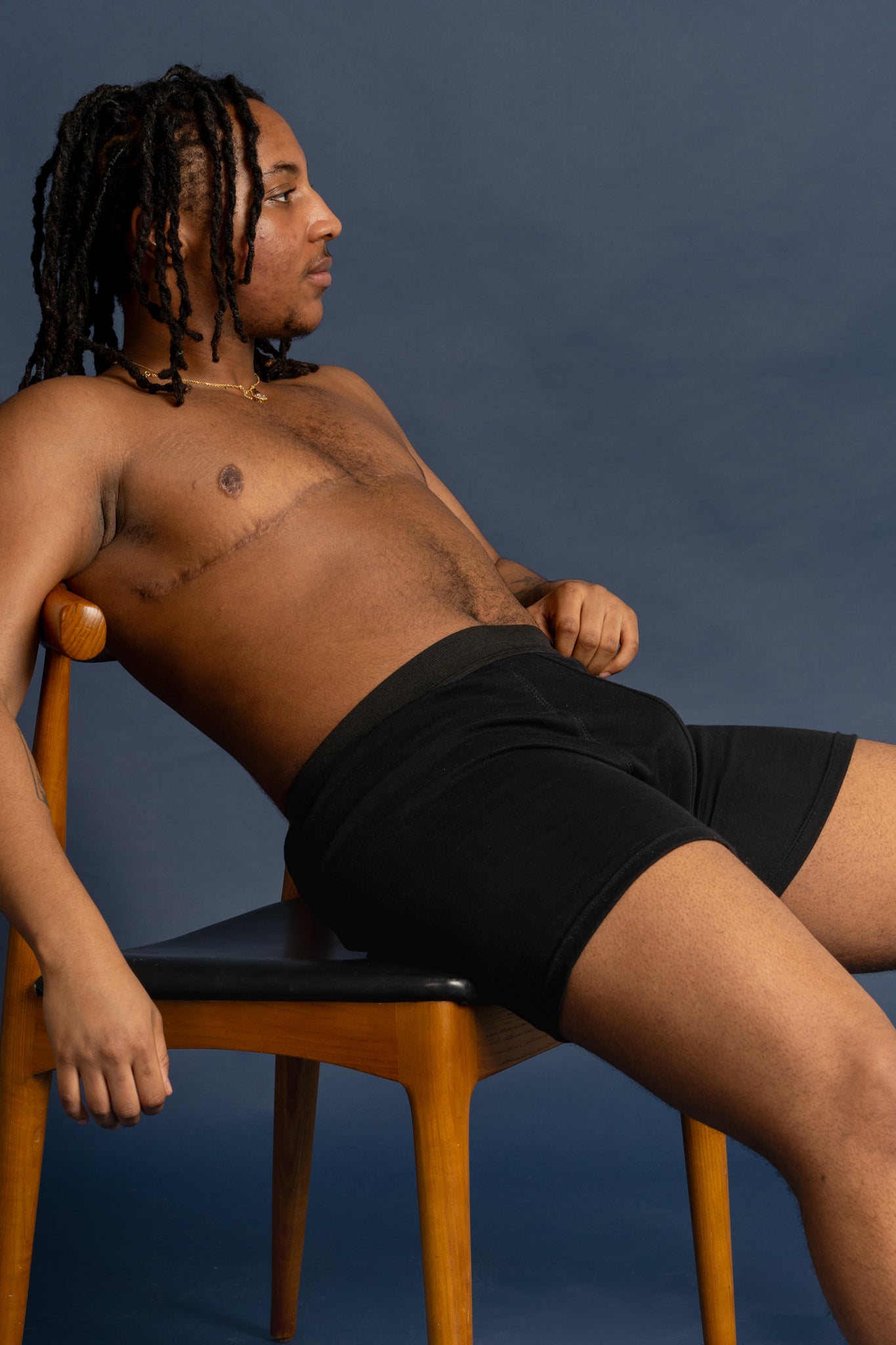 A shirtless trans man sitting on a chair wearing spectrum outfitters boxers