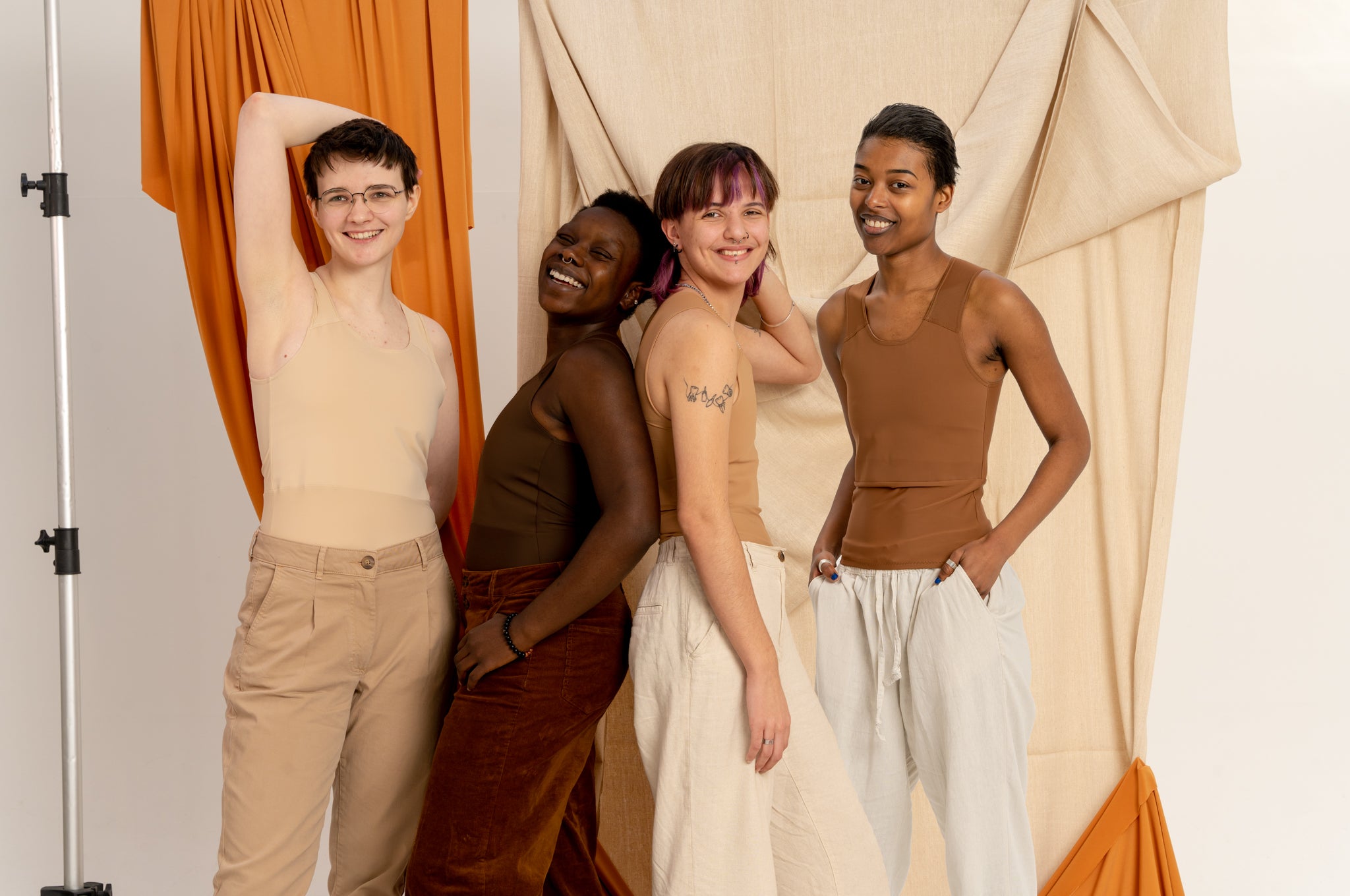 Four people posing together wearing 4 different skin tone chest binders in front of orange and beige curtains