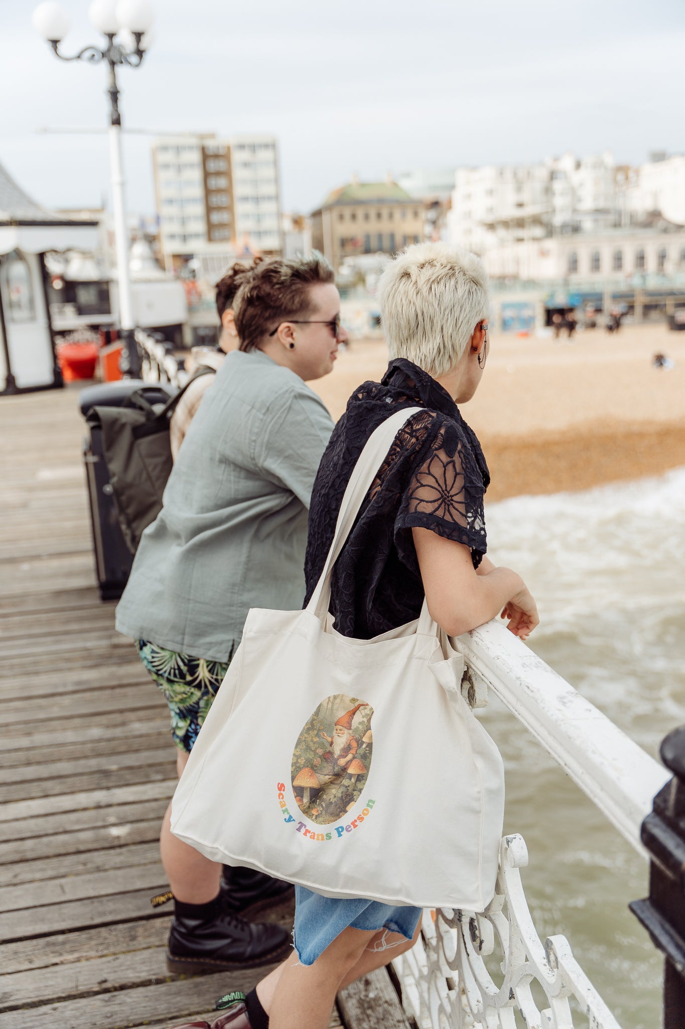 Two people standing on a pier with one holding a tote bag with a woodland gnome graphic reading 'Scary Trans Person'