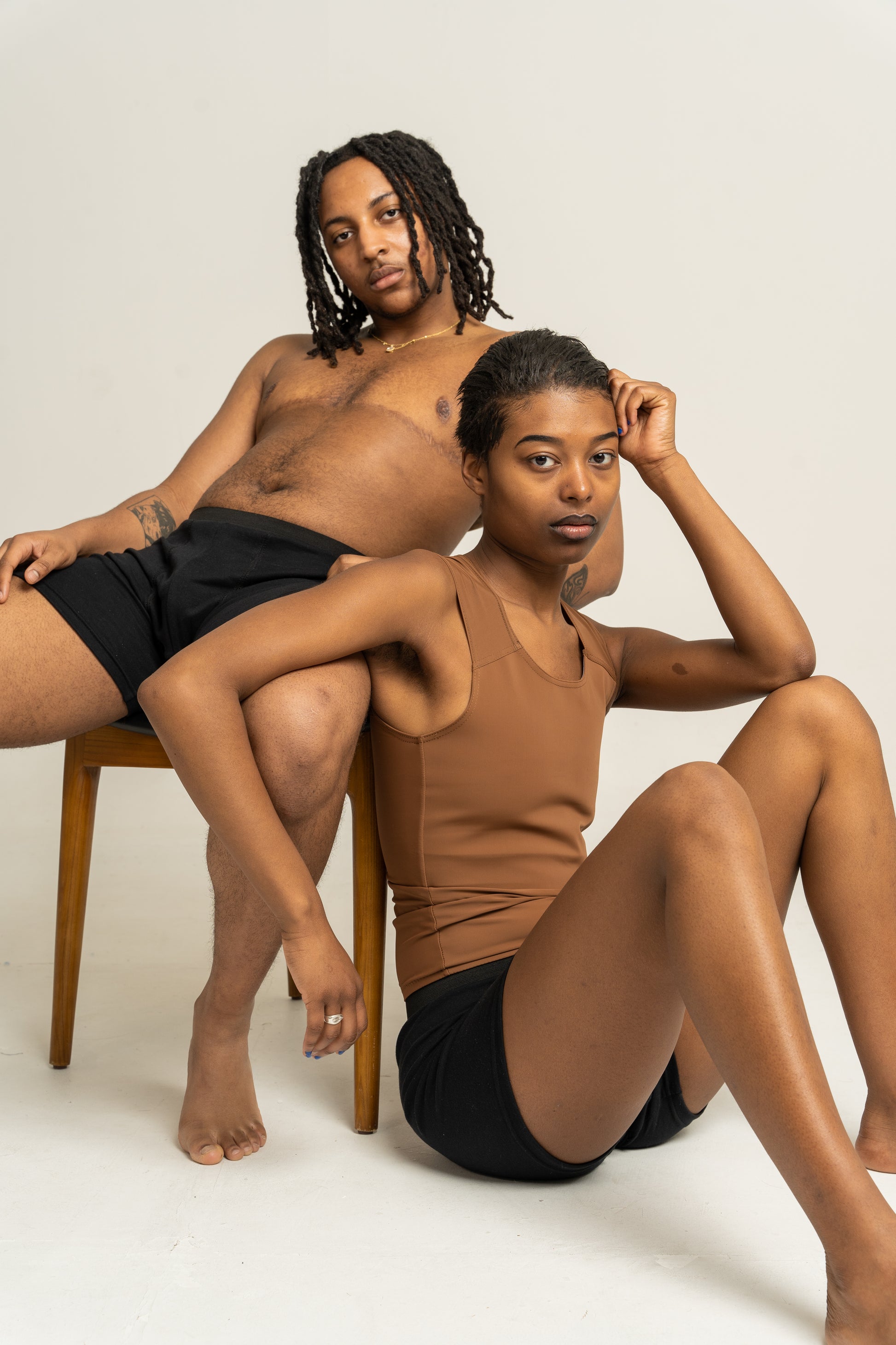 Two people sitting on a white background, one wearing a brown chest binder top and black boxers, the other in black boxers.