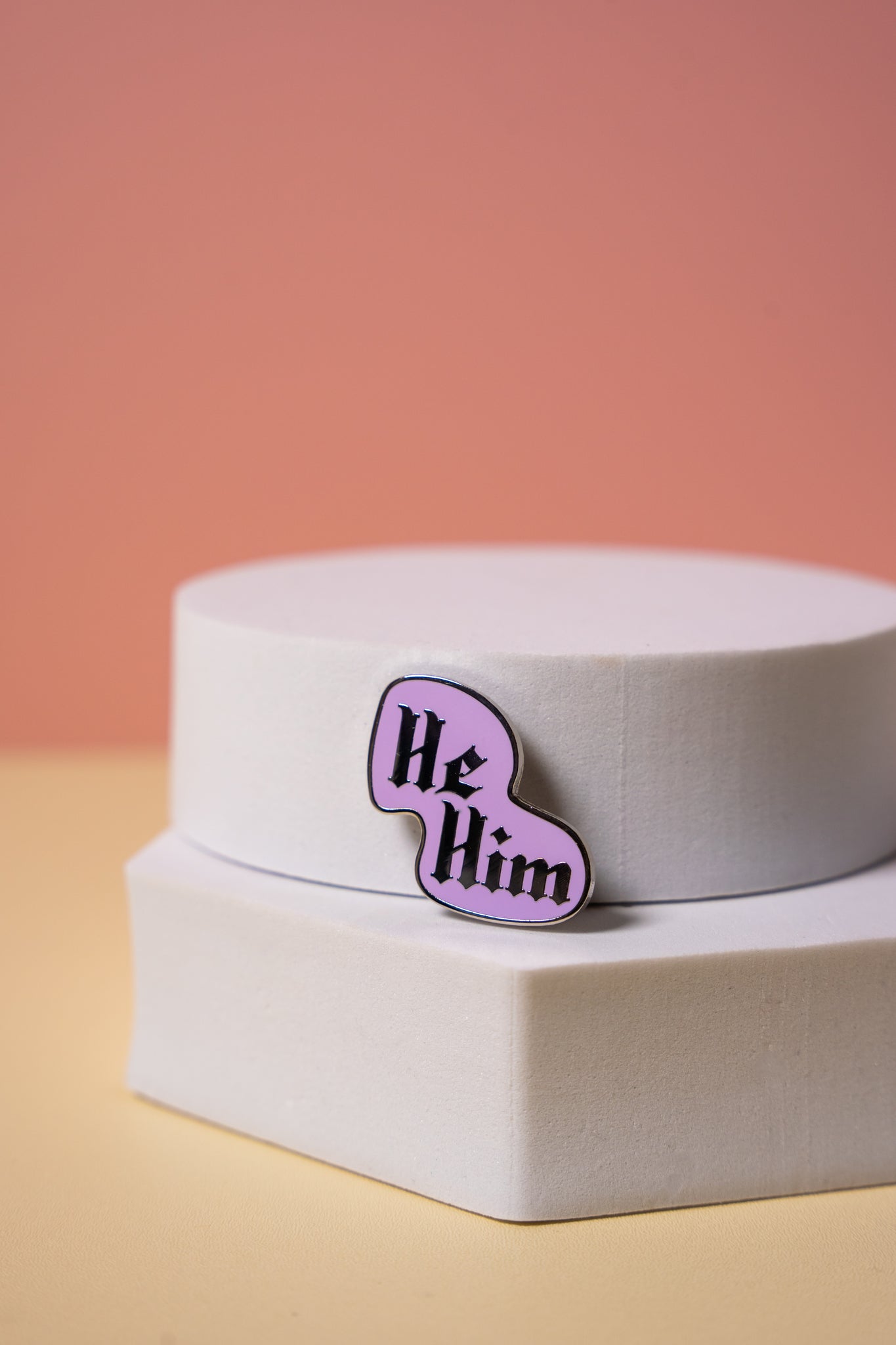 Purple gothic style 'He/Him' pronoun pin on a white stand with a pink background