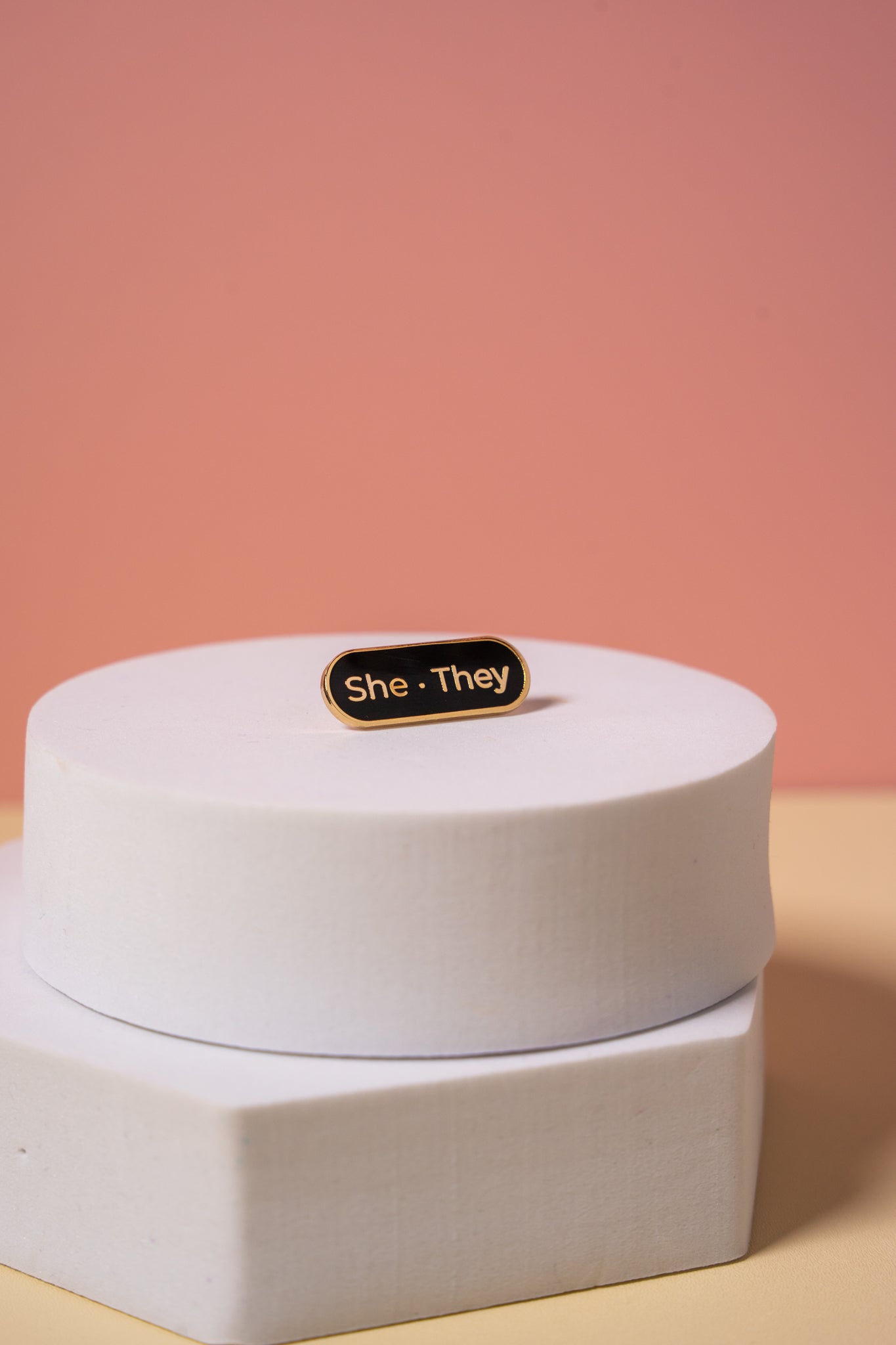 A gold and black enamel  'She They' pronoun pin on a peach background