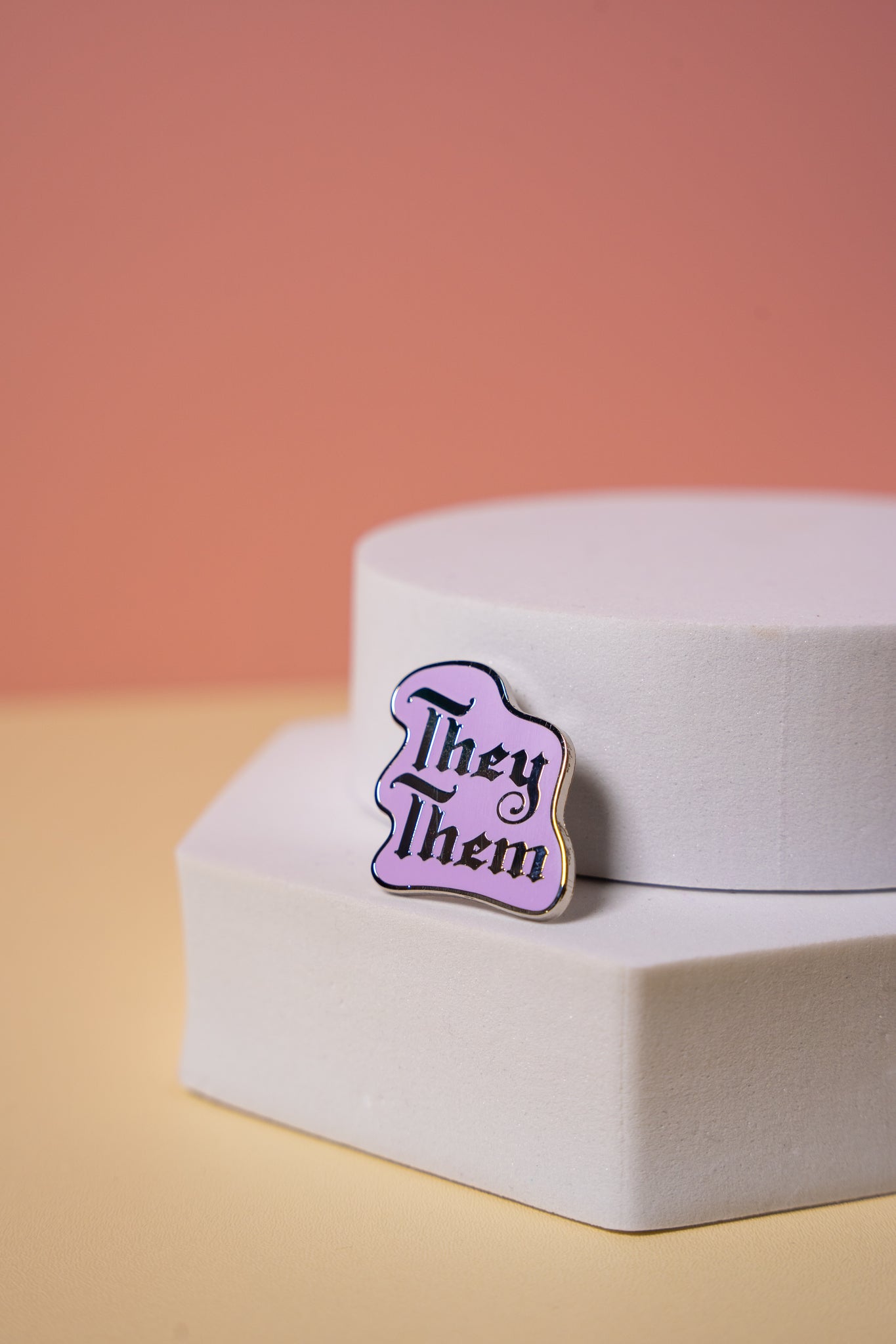 Enamel pin with 'They/Them' text on a white stand against a peach background