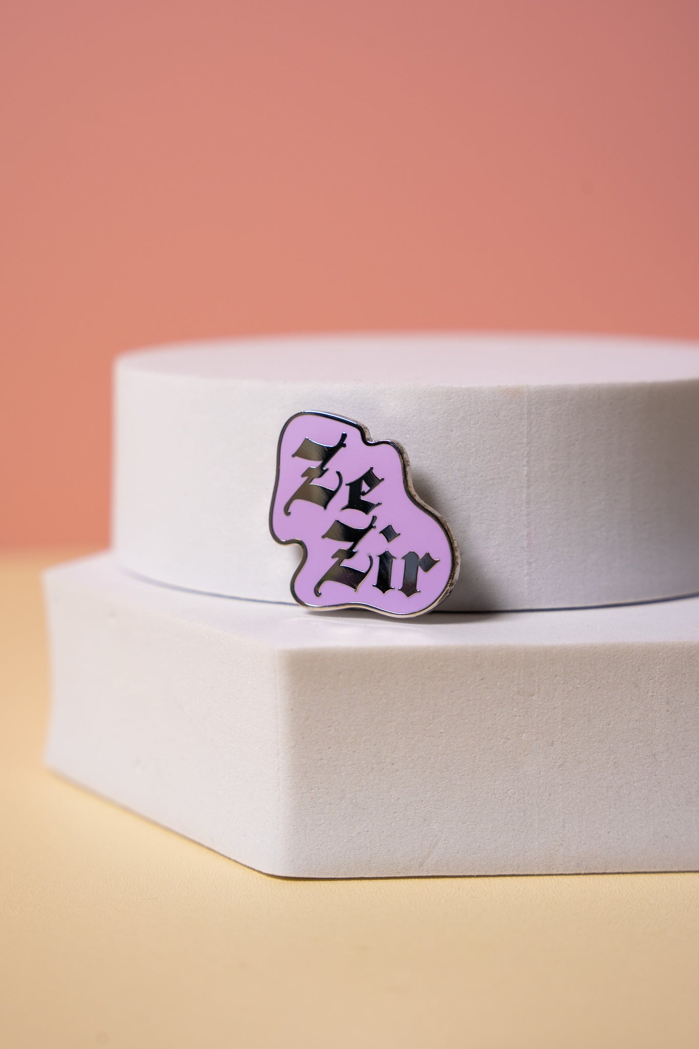 Purple enamel pin with 'Ze Zir' text on a white hexagonal base against a pink background
