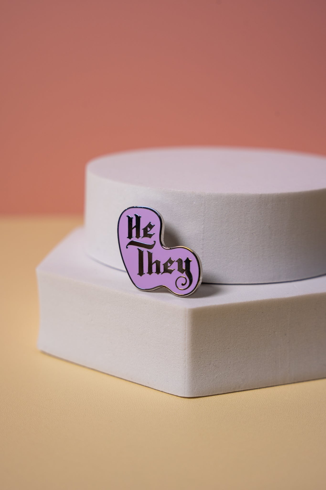 Purple enamel pin with 'He/They' text on a white stand against a gradient background