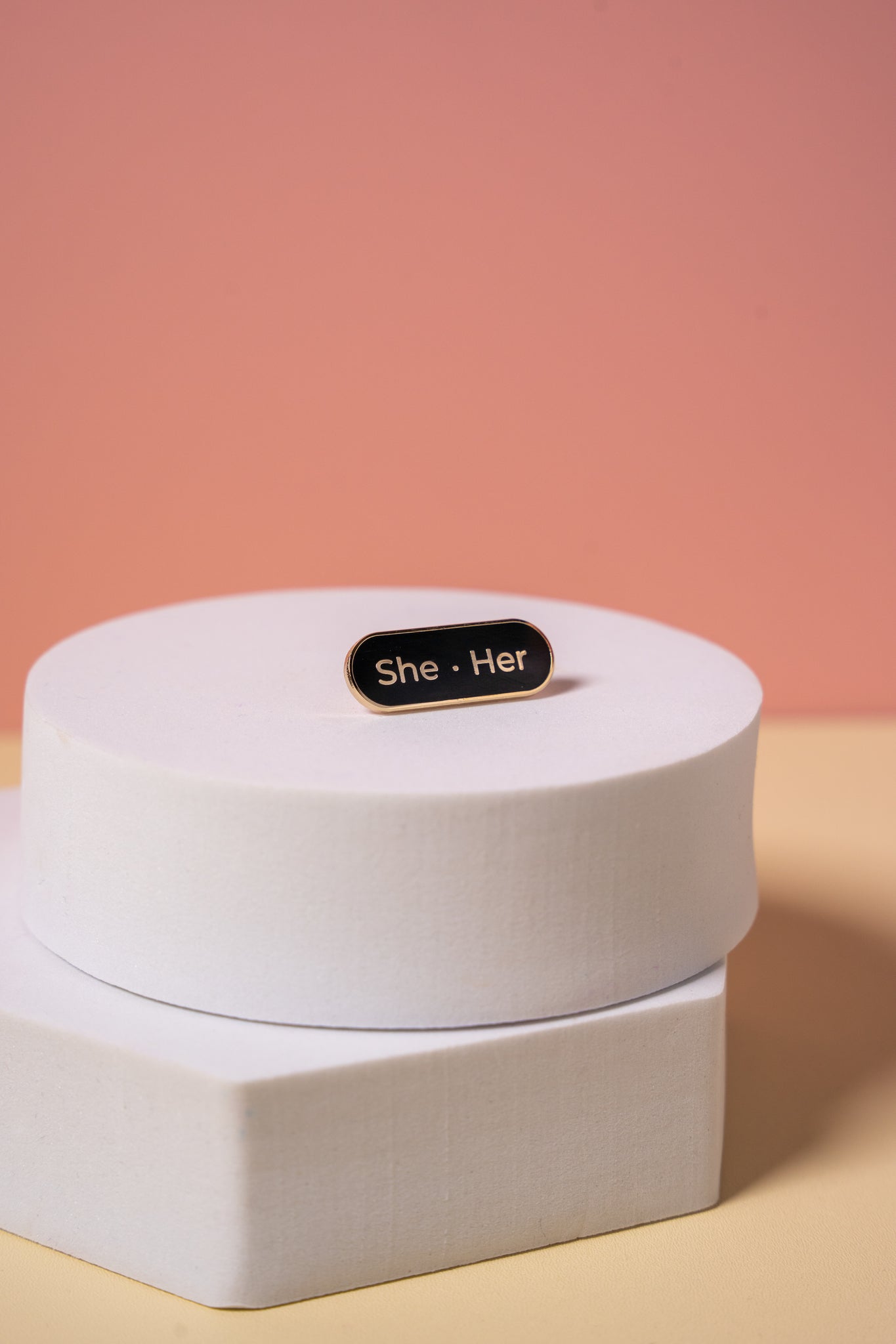 a black enamel 'She-Her' pronoun pin on a white box with a pink gradient background