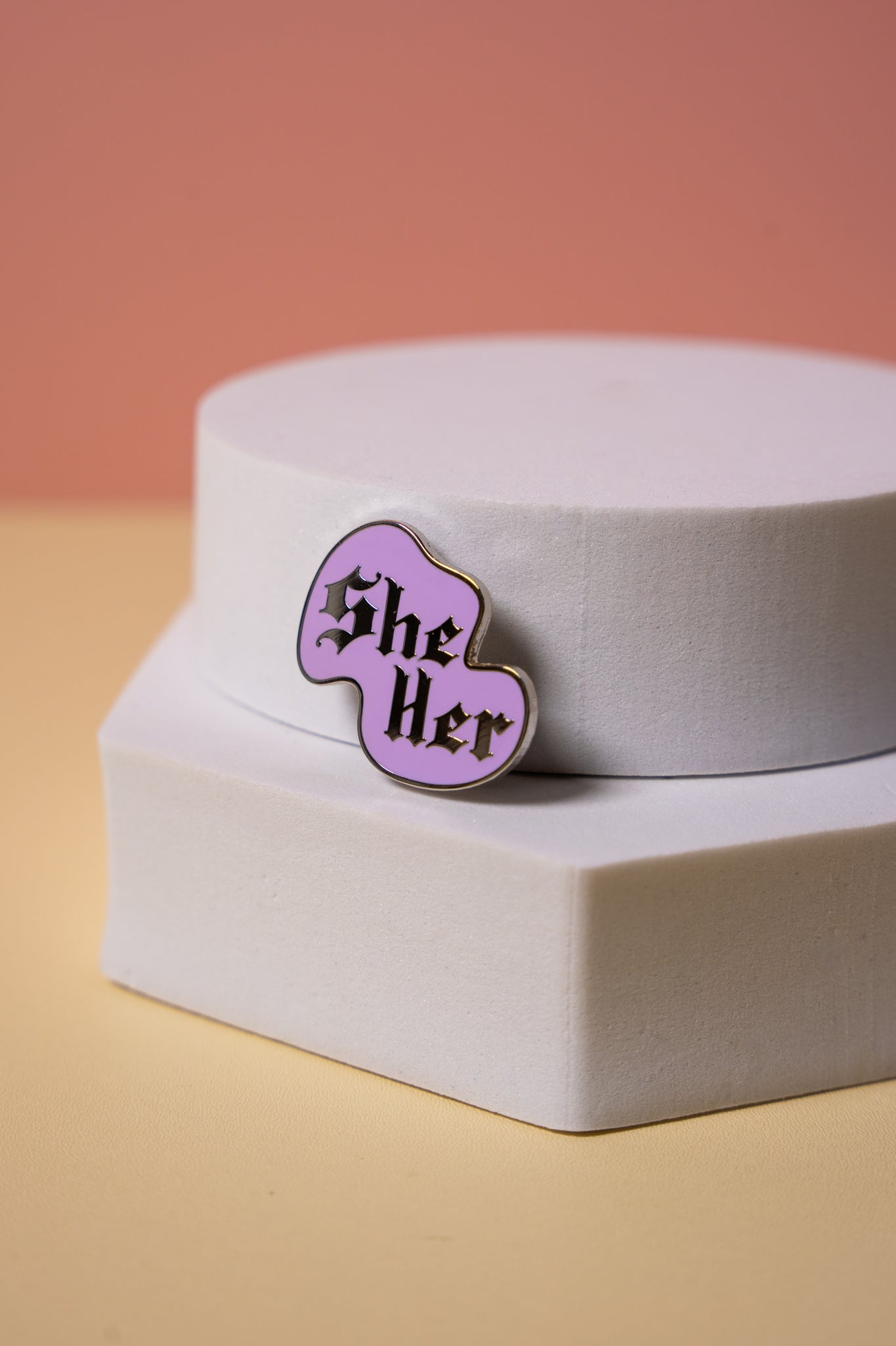 Purple 'She Her' enamel pin on a white box with a pinkish-orange background