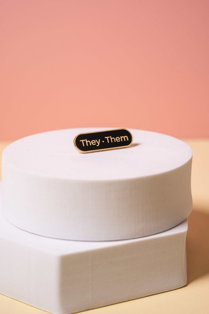 A gold and black enamel  'They Them' pronoun pin on a peach background